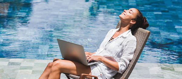 Woman working on laptop while basking in the sun with a pool behind her